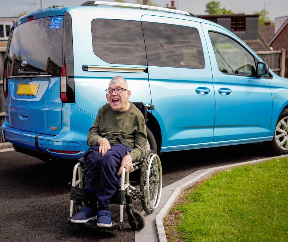Un homme en fauteuil roulant sourit près d'un véhicule bleu adapté aux personnes à mobilité réduite (TPMR), garé dans une allée. On aperçoit une pelouse et des maisons en arrière-plan.
