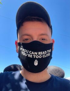 Ross wearing a black facemask with white writing saying, "If you can read this, you're too close"