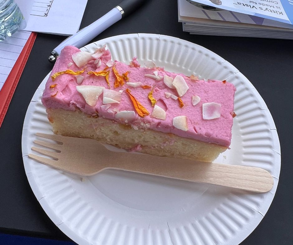 Slice of free dragon fruit cake with pink frosting topped with dried flower petals, served on a white paper plate with a wooden fork on a black table at Allied Mobility's event held at the Historic Dockyard Chatham