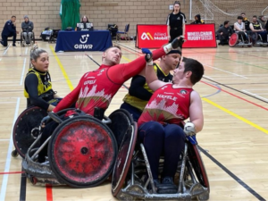 Two male wheelchair rugby players in red vests with hands up in the air to block male wheelchair ruby player in black vest shielding the ball. Female wheelchair rugby player in black vest looks on. 