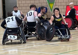 Three male wheelchair rugby players in white vests and two male and one female wheelchair rugby players in black vests challenging for the ball. 