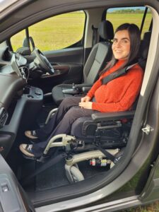 Emma, wears an orange jumper and travels in her car in the upfront position in her powerchair 