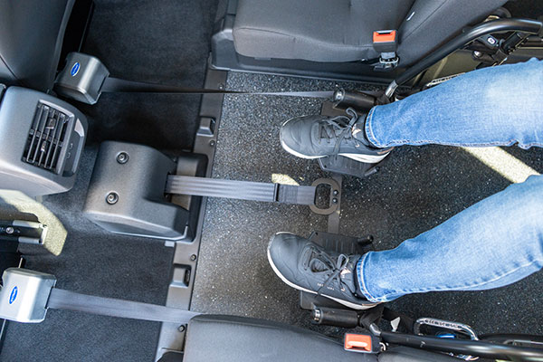 Person wearing jeans and black trainers uses a foot-operated accelerator pedal in a vehicle, with one foot on the pedal and the other resting on the floor between two car seats.