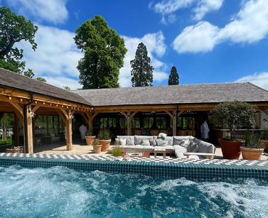 Open air, wood canopy with slated grey roof. White couch in front of this and blue swimming pool at forefront of image.