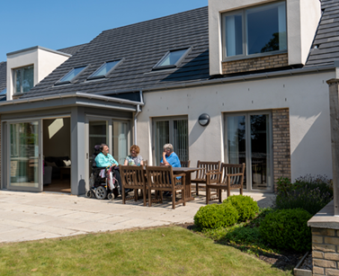 Three ladies in distance drinking coffee at an outdoor wooden table in the sunshine. Kitty is sitting in her powerchair in a light blue top.