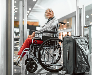 Older female is smiling from her wheelchair with grey hair, grey and white striped jumper and red trousers. Her black suitcase is next to her.