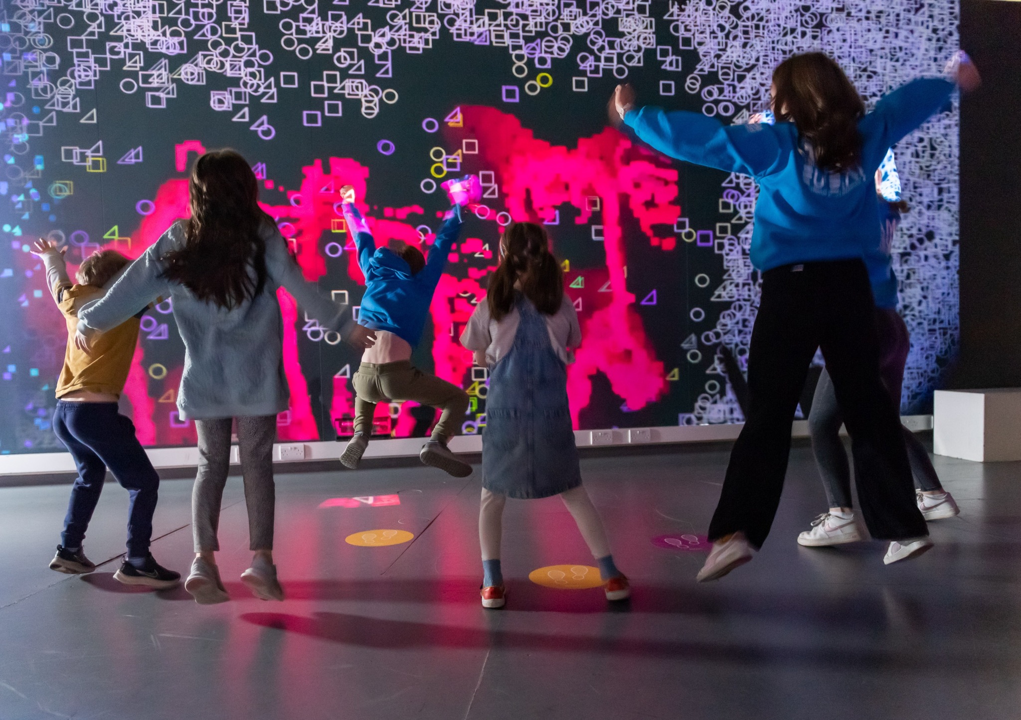 Five children of various ages star jump in front of a light exhibit at Eureka! The National Children's Museum