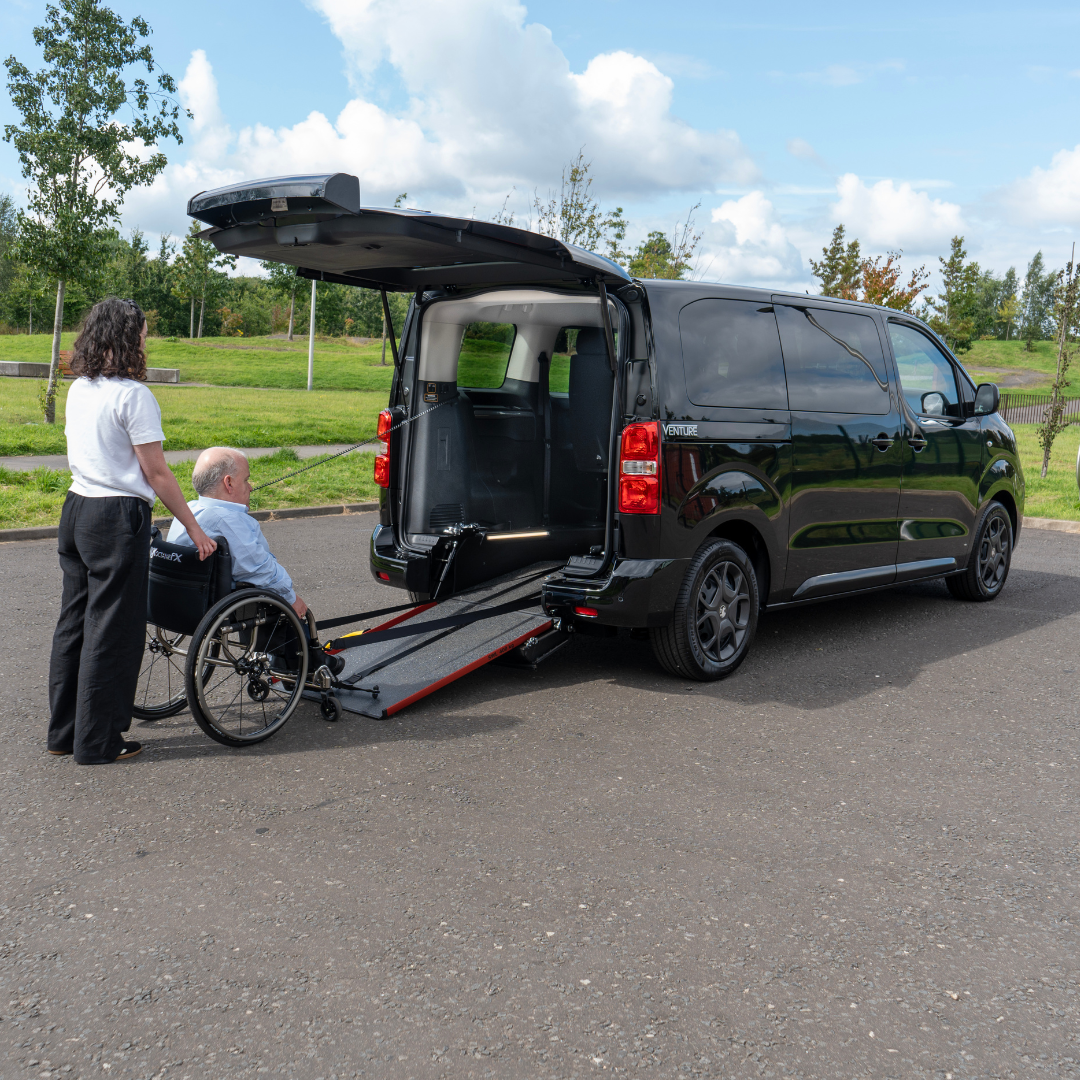 A man in a wheelchair is positioned at the bottom of the ramp of a Vauxhall Venture wheelchair accessible vehicle. A woman stands behind him for support.