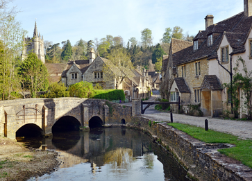 A tranquil Cotswolds village with traditional stone houses, a small stone bridge crossing a calm river, and a church with a tall spire in the background, surrounded by lush greenery.