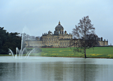 Castle Howard House and Gardens with a large central dome and smaller spires, surrounded by green lawns and trees, with fountains spraying water in the foreground.