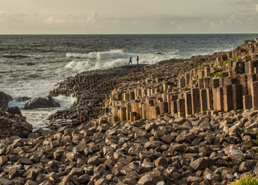 The Giant's Causeway in Northern Ireland, featuring interlocking hexagonal basalt columns formed by volcanic activity, with two people walking along the rocks and waves crashing in the background.