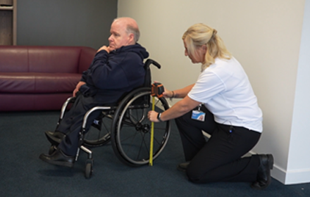 An woman with blonde hair in Allied Mobility uniform measures the wheelchair of a man in a blue jumper.