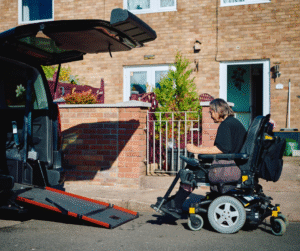 Arlene in her powerchair positioned at the bottom of the ramp of her accessible vehicle. The scene is outside a brick house with a small front garden, metal railing, and an open front door. The wheelchair has large rear wheels, a cushioned seat, and a backpack attached to the back. The ramp leads into the vehicle, designed for wheelchair access. Bright daylight illuminates the setting.