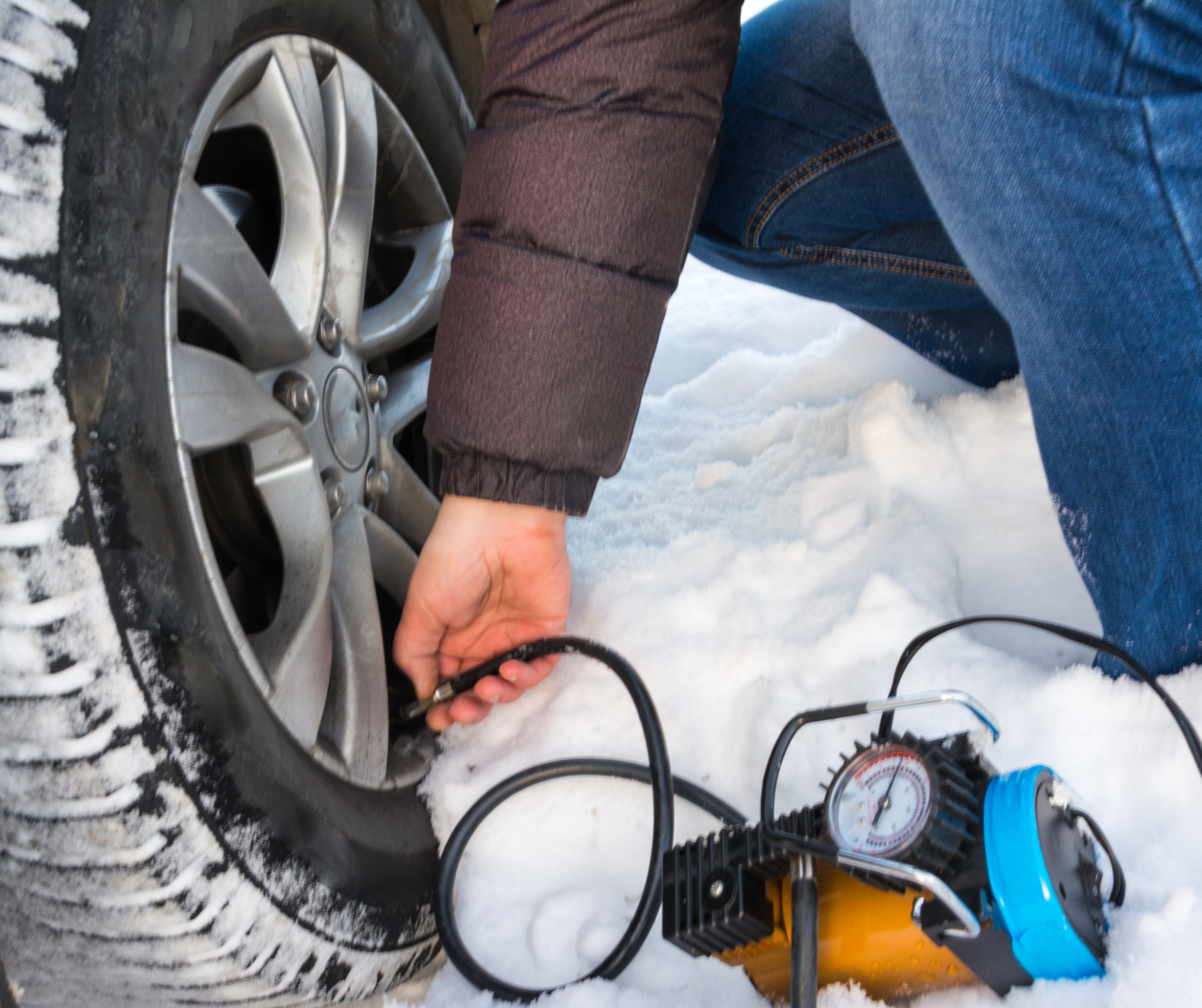 Person inflating a car tire with a portable air compressor on snowy ground, focusing on the wheel and equipment for winter car maintenance.