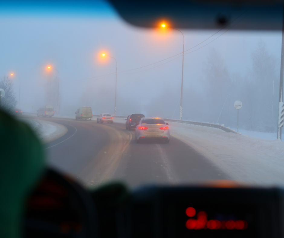 Cars drive on a snowy, curved road in dense fog with limited visibility. Streetlights cast a warm glow. Trees and road signs are faintly visible in the background.