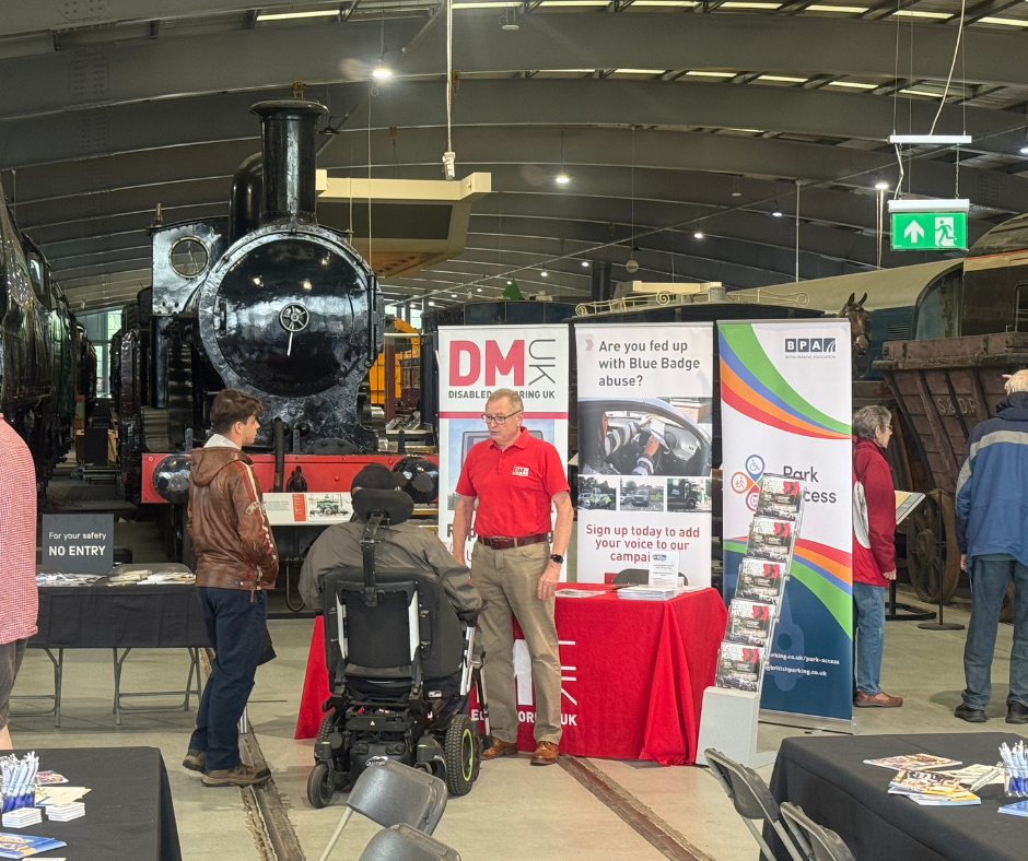 An indoor exhibition with a historic black steam locomotive in the background. In the foreground, a stall displays banners for Disabled Motoring UK and campaigns against Blue Badge abuse. A person in a powered wheelchair is speaking with a representative at the stand, while others browse nearby tables.