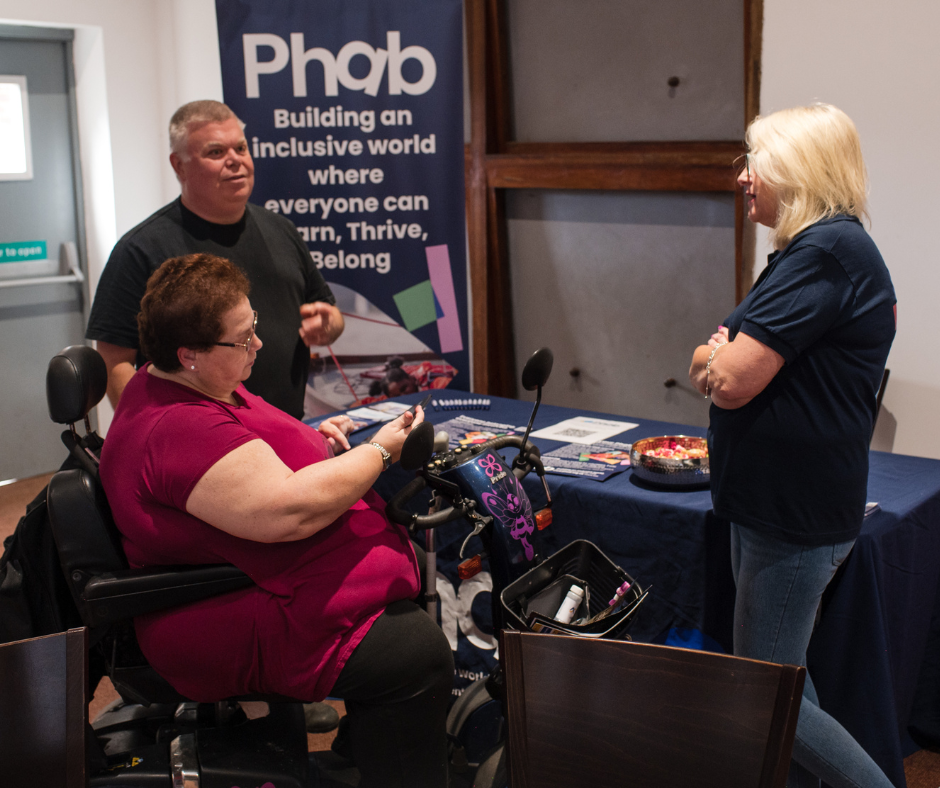 A community information stand for Phab, featuring a banner that reads “Building an inclusive world where everyone can learn, thrive, belong.” A person using a mobility scooter is engaged in conversation with two individuals at the table, which is covered with leaflets and a bowl of sweets.