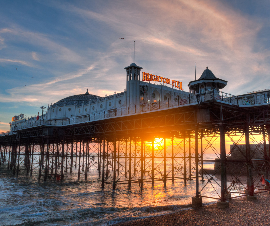 A view of Brighton Pier at sunset, with the sun shining through the pier’s metal supports and illuminating the sky with warm orange and blue tones.
