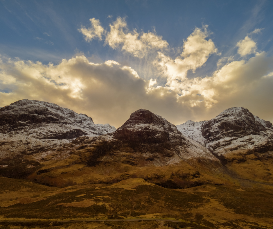 A dramatic mountain landscape in Glencoe, with snow‑dusted peaks under a glowing sky filled with bright clouds and soft sunlight.