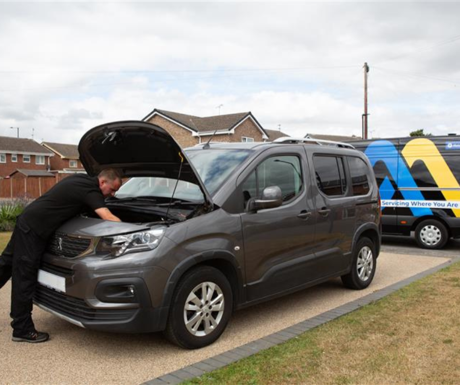 A grey WAV with its hood open being inspected by a mechanic in a residential area. A service van with a blue and yellow design is parked nearby.