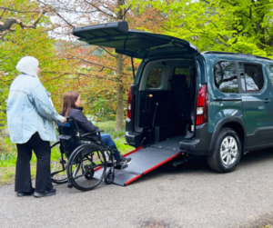 A green wheelchair-accessible vehicle with its rear door open and ramp extended, parked on a road surrounded by trees. A person is pushing a wheelchair user toward the ramp.