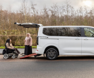 A white wheelchair-accessible vehicle with the word “Independence” on the side, rear door open and ramp extended, parked on a roadside. Two people are positioned near the ramp, one seated in a powered wheelchair.