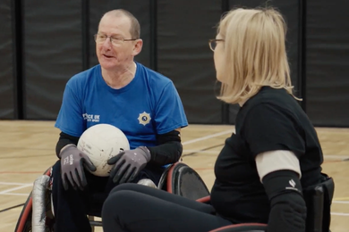 Tony chatting to a fellow wheelchair rugby athlete in their wheelchair rugby chairs. He holds a ball in his lap.