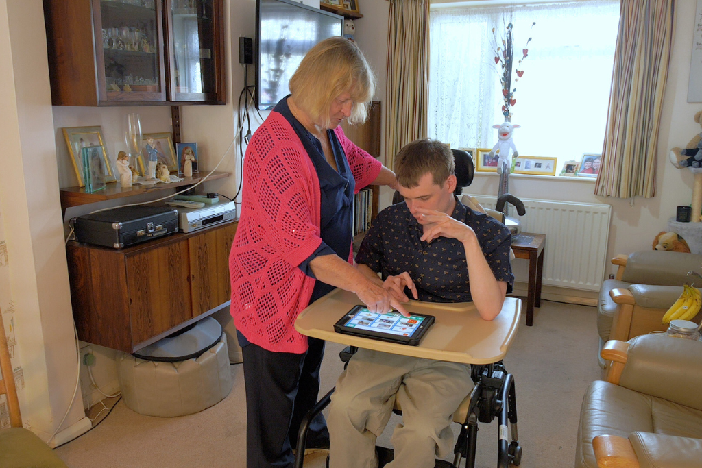 Caregiver, Carol, assists Charles with a tablet device in a cozy living room setting. Charle's wheelchair has a tray holding the tablet, and Carol is pointing at the screen to provide guidance. The room features a wooden cabinet with decorative items, a television on the wall, framed photos on the windowsill and comfortable armchairs.