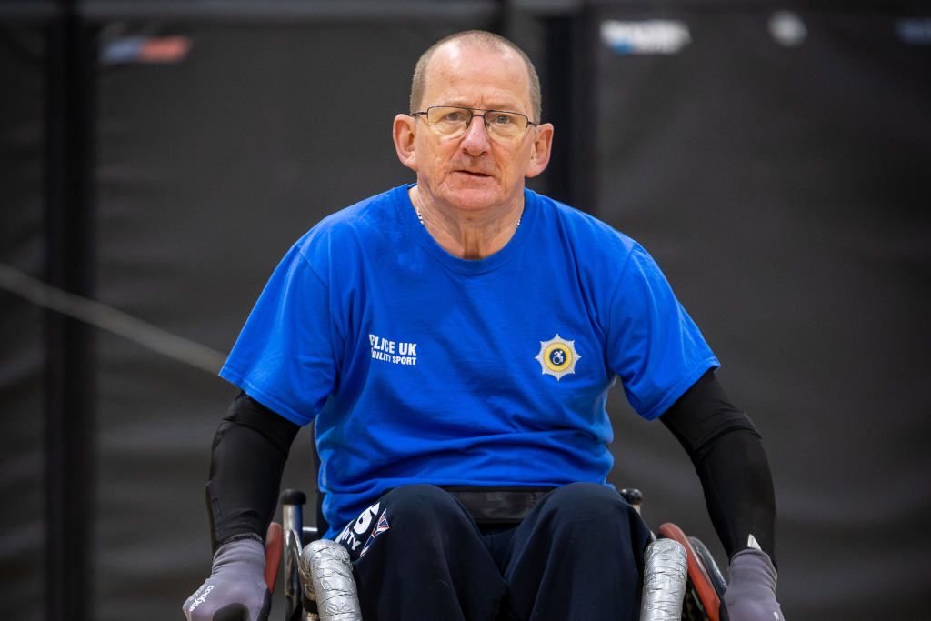 Tony in his wheelchair wearing a bright blue T-shirt with a logo and text that reads ‘SLK UK Health Sport.’ Tony has black sleeves under the T-shirt and is positioned indoors against a dark background.