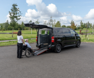 A black wheelchair-accessible vehicle with its rear door open and ramp extended, parked on a paved area near a grassy park. A person is assisting another individual in a wheelchair toward the ramp.