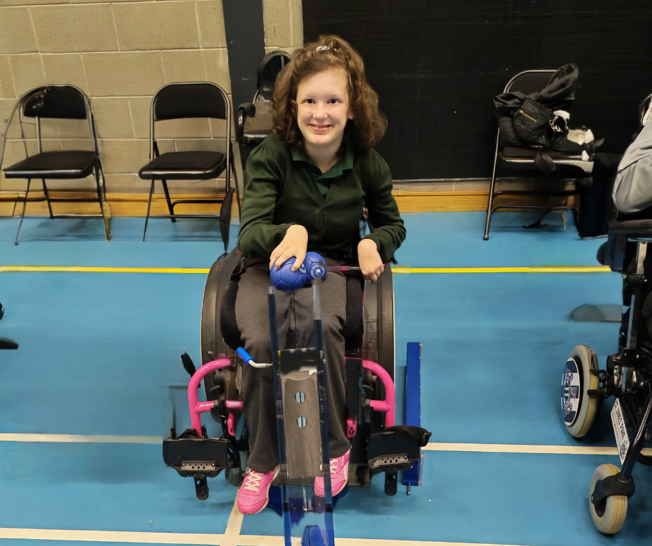Lana seated in a wheelchair on a sports court, holding a blue ball positioned in front of a ramp used for boccia. The wheelchair has bright pink accents, and there are chairs and another wheelchair visible in the background.