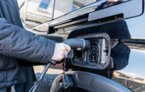 A person wearing protective gloves lifts a car battery out of an engine compartment, holding it by the built‑in handles while the car’s hood is open.
