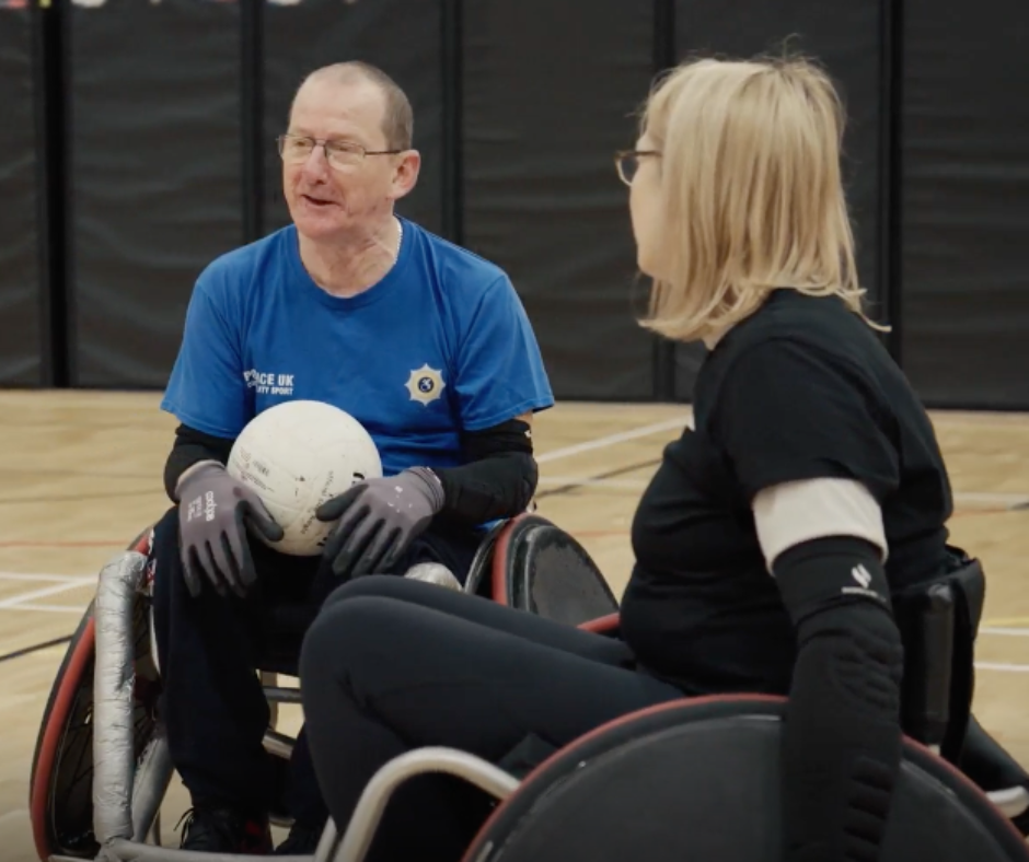 Tony chatting to a fellow wheelchair rugby athlete in their wheelchair rugby chairs. He holds a ball in his lap.