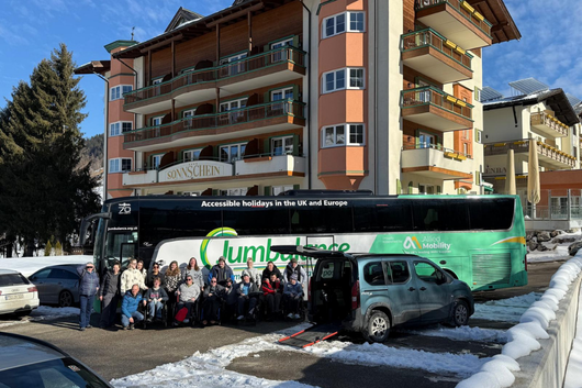 A large Jumbulance accessible coach parked outside a multi-storey hotel in a snowy mountain location, with a group of travellers and volunteers gathered in front of it. A wheelchair-accessible vehicle with a ramp lowered is also visible.