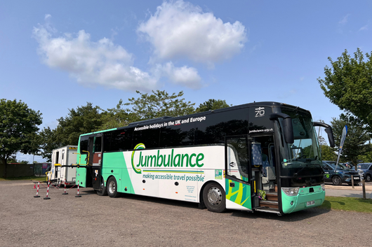A Jumbulance accessible coach parked on a sunny outdoor site, with clear blue sky and trees in the background. The side of the coach displays the Jumbulance branding and the slogan “making accessible travel possible.