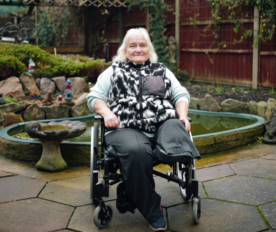 A woman sitting in a wheelchair in a garden area with a small pond and decorative stone features behind her.