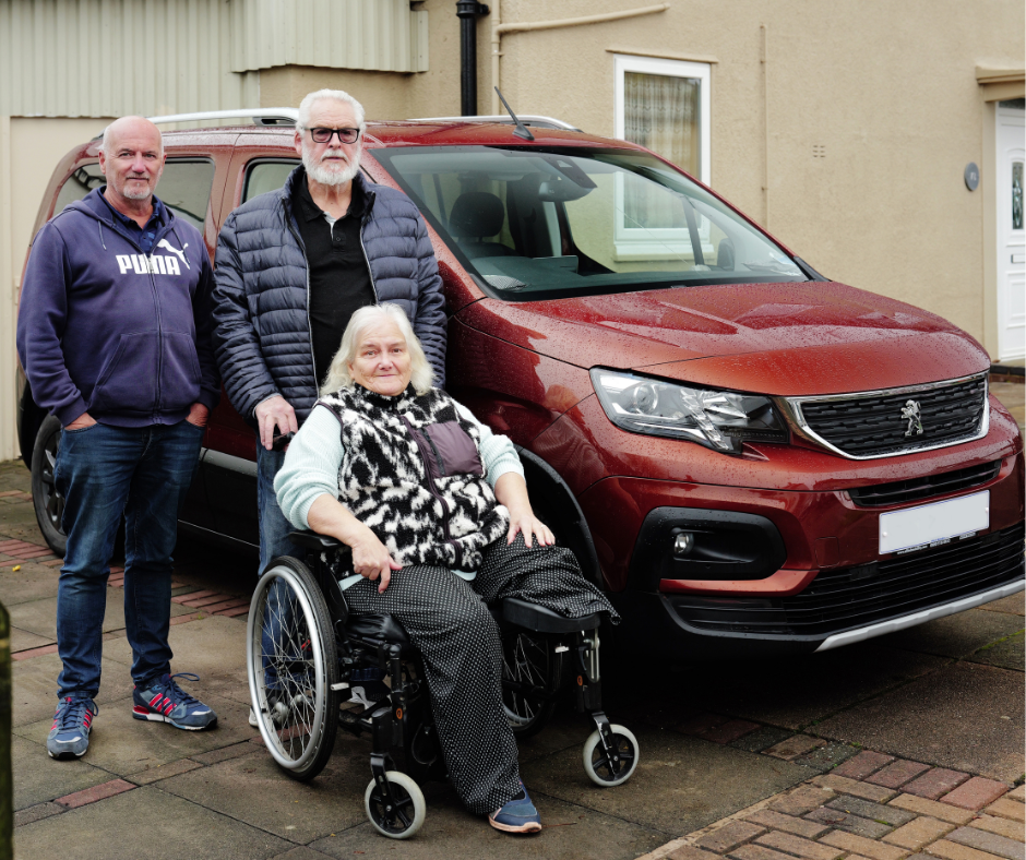 A woman in a wheelchair on a driveway with two men standing beside her, positioned in front of a red Peugeot wheelchair accessible vehicle.
