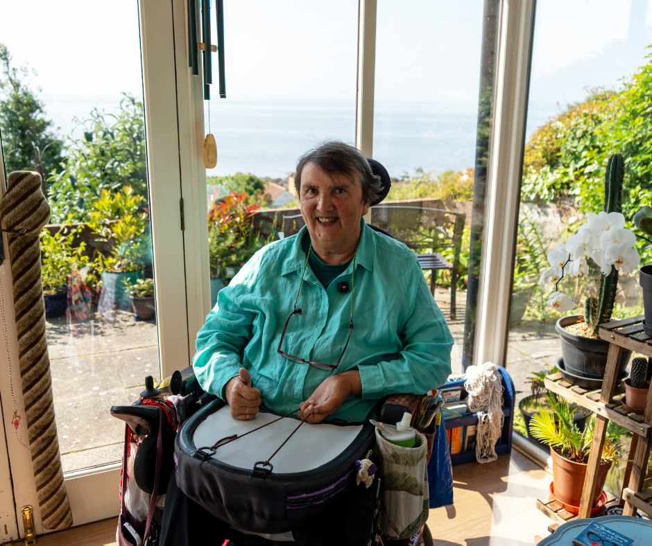 A person seated in a wheelchair is indoors near large glass doors overlooking a sunny garden and a distant view of the sea. They are wearing a turquoise shirt and have a small drum resting on their lap with one hand positioned on it. The room around them contains various plants, shelves, and natural light streaming in from outside.