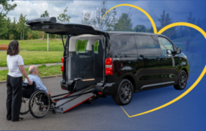 A person wearing protective gloves lifts a car battery out of an engine compartment, holding it by the built‑in handles while the car’s hood is open.