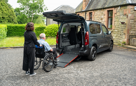 A woman helps a man in a wheelchair up a ramp into the rear of a wheelchair accessible vehicle parked outside a stone building on a sunny day, showcasing the accessibility and ease of use provided by WAVs.