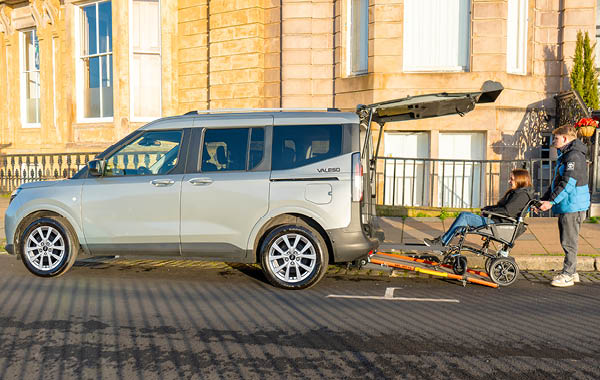 Side view of a grey Ford Valeso wheelchair accessible vehicle parked on a city street with the rear access ramp extended and a wheelchair user being assisted towards the vehicle.