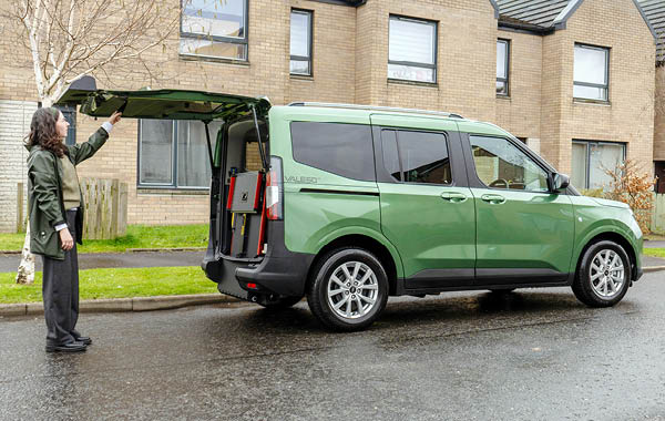 Side‑rear view of a green Ford Valeso wheelchair accessible vehicle with the tailgate raised and rear access ramp stored upright.