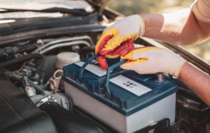 A person wearing protective gloves lifts a car battery out of an engine compartment, holding it by the built‑in handles while the car’s hood is open.