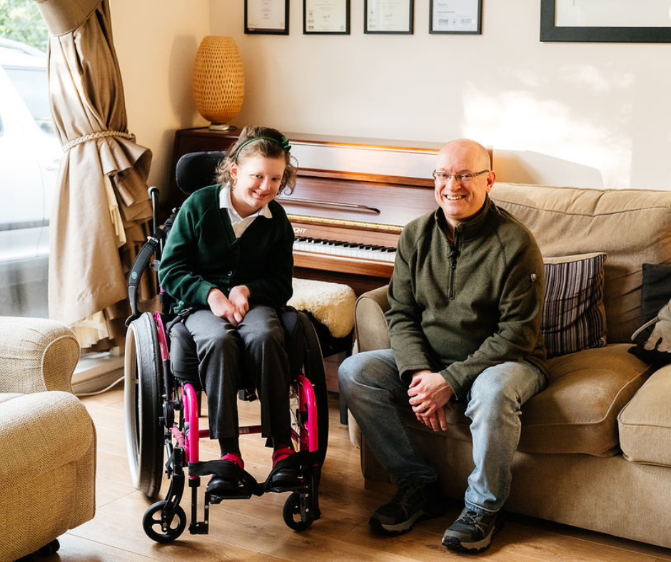 Lana in a pink‑framed wheelchair sits next to her dad, Paul, who is seated on a sofa in a warmly lit living room. A piano stands behind them with framed certificates on the wall above it. The room features soft furnishings, including armchairs and cushions, creating a comfortable, homely setting.