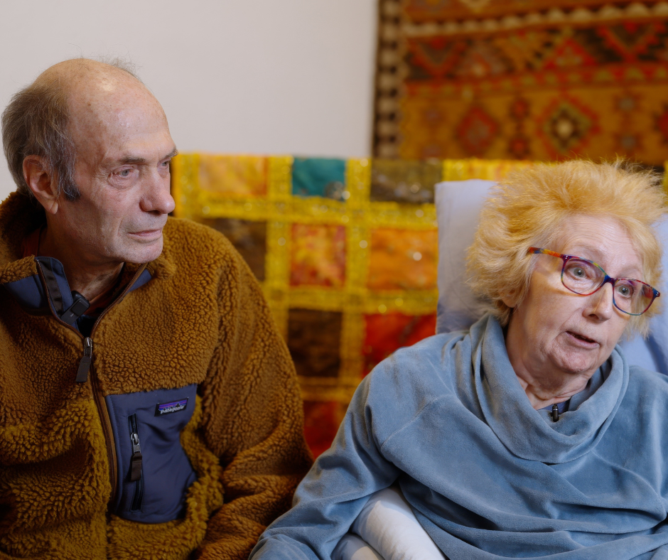 Frances and Dale sit closely together indoors. Dale is wearing a brown fleece jacket, and Frances is dressed in a light blue robe while resting against pillows. Behind them is a colourful patchwork quilt and a patterned tapestry hanging on the wall.