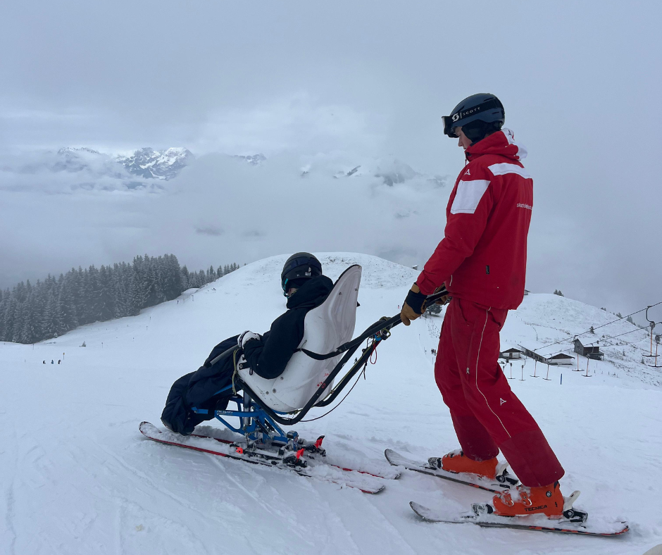 An instructor in a red ski uniform guides a sit‑ski down a snowy mountain. The sit‑ski rider is securely seated, with snow-covered hills and cloudy mountain scenery in the background.