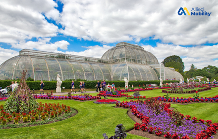 Victorian glasshouse surrounded by colorful flower beds, green lawns, and visitors walking in a botanical garden under a partly cloudy sky.