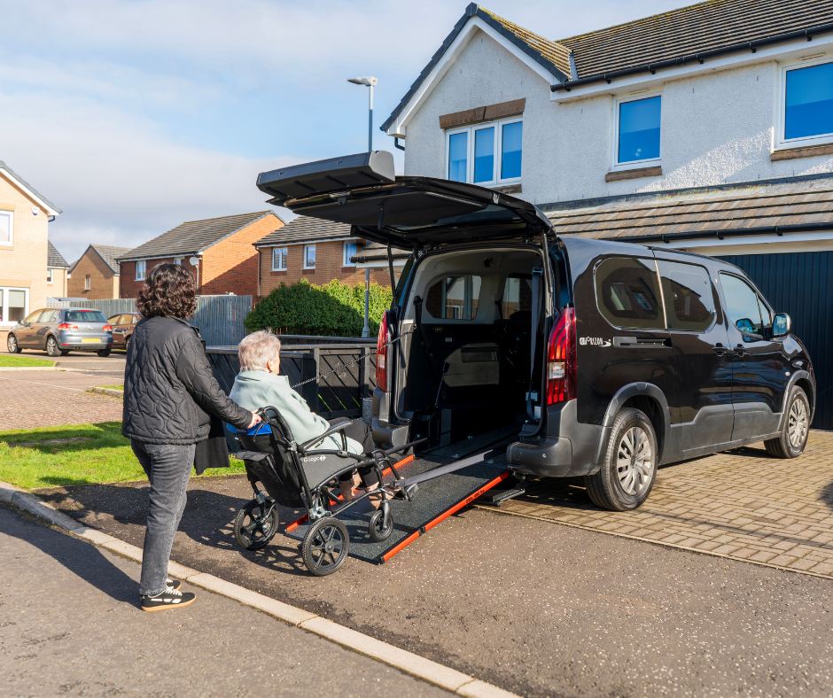 A woman helps a female wheelchair user up the ramp of a wheelchair accessible vehicle which is parked outside a suburban house.