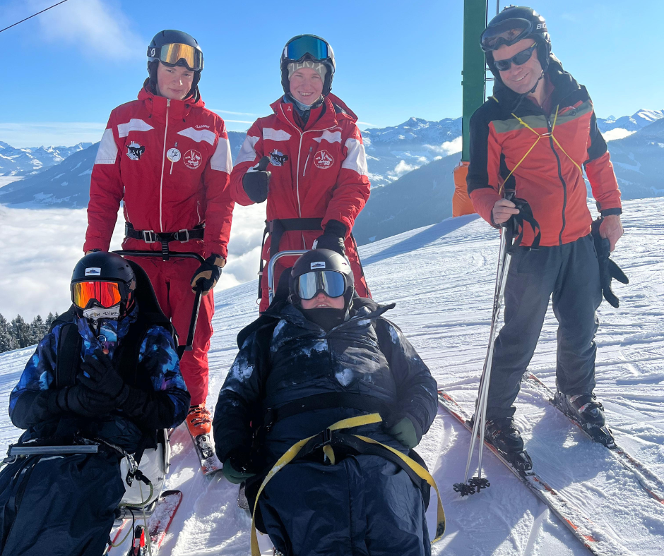 A group of adaptive skiers and instructors stand together on a snowy mountain slope. Two participants are seated in sit‑skis while instructors in red ski uniforms stand beside them, with a backdrop of clear blue sky and mountain peaks.