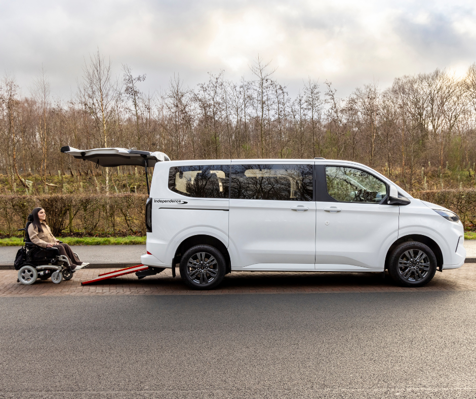 A wheelchair user waits behind a white accessible vehicle with its rear ramp extended, parked beside a road with trees in the background.